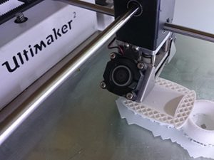 The image shows a desktop 3D printer, an Ultimaker, in the process of 3D printing using hot extruded plastic filament which is used to outline and fill in each x,y profile of the 'sliced' model, to produce a three dimensional object from the 3D digital model's data. 