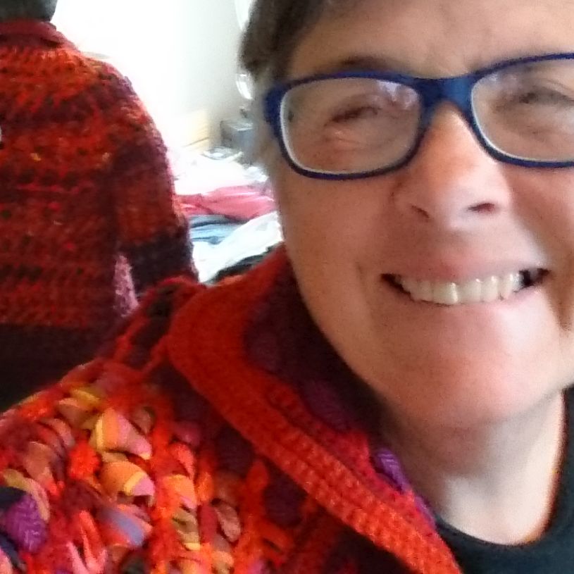 Image of Ann Marie Shillito wearing a jacket. crocheted from waste red/orange carpet wool and interwoven with strips of waste and scrap fabrics left over from making her own clothes. She made this in 1979/80 and is now on another journey: how 3D digital technologies support a designer/maker's more environmentally responsible practice.